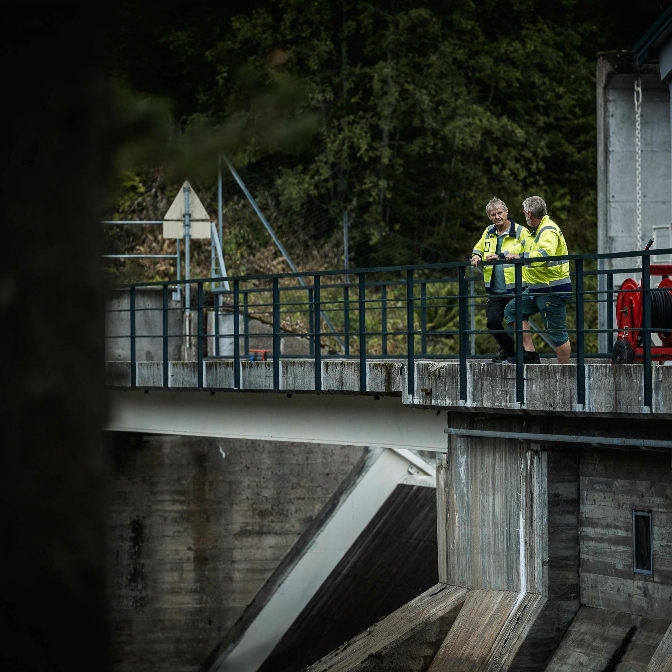 Foto: To personer iført arbeidstøy som står ved rekkverk og ser ned fra et vannkraftverk