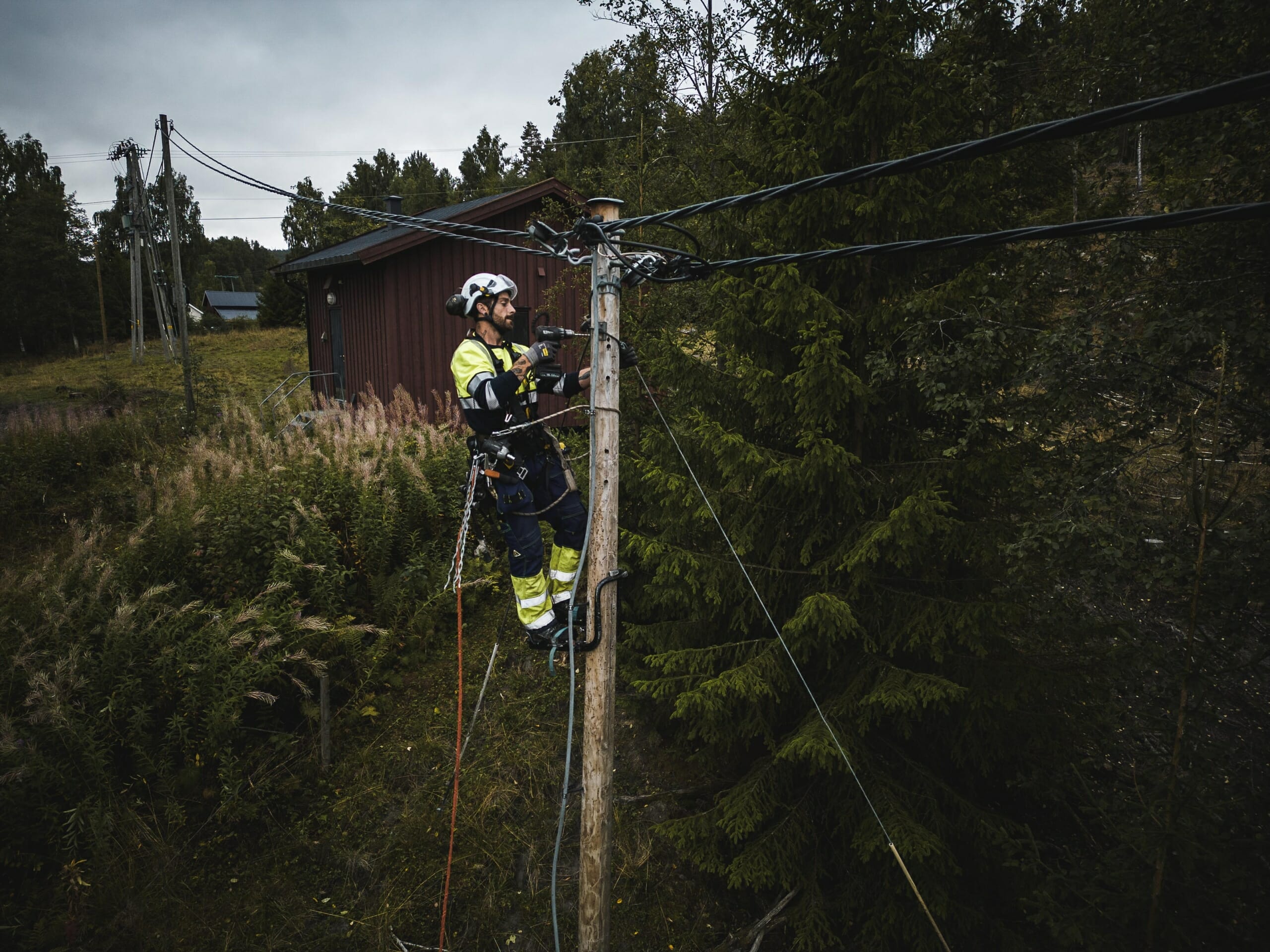 Foto: Person i fullt arbeidsutstyr monterer fiberkabel på en stolpe i et landlig område.