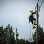 Foto: Person i fullt arbeidstøy, sikret og i toppen av en elektrisk stolpe og monterer en kabel.