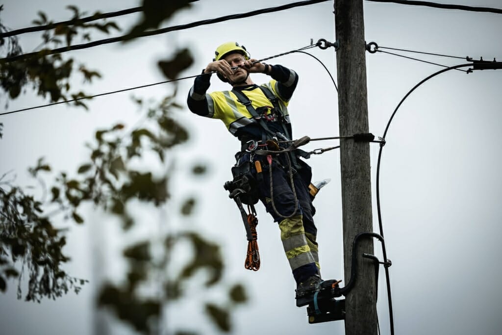 Foto: Person i fullt arbeidstøy som er festet til en elektrisk stolpe og monterer en kabel.